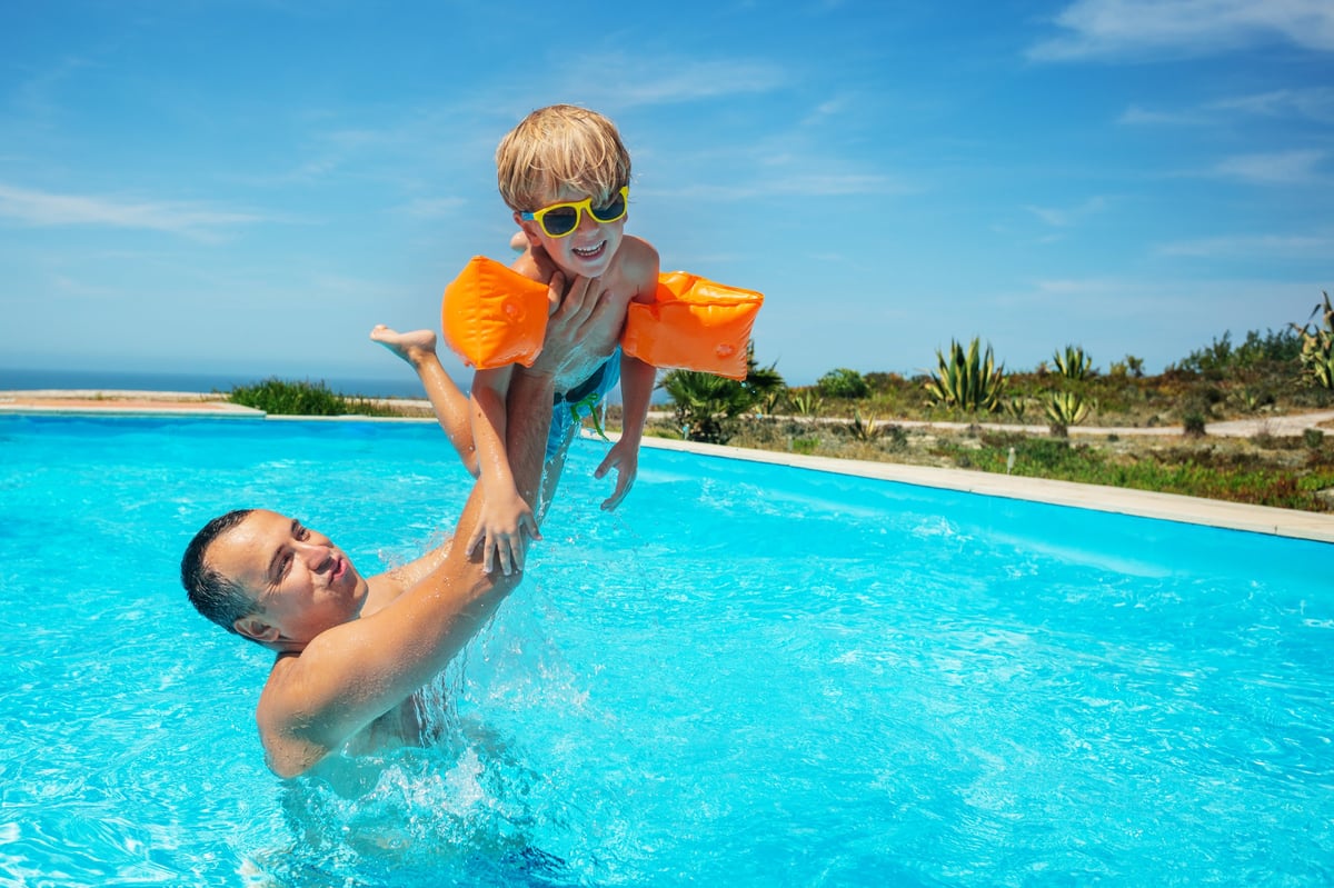 In swimming pool adult lifting a child high above the water in a playful manner, boy with sunglasses wearing floaties on their arms to aid swim