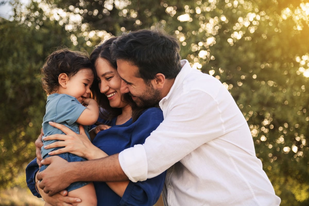 Una familia feliz con papá, mamá e hijo pequeño abrazándose y sonriendo en un entorno natural con césped y árboles