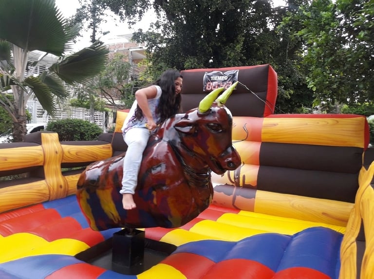 Child riding a mechanical bull on a colorful inflatable bounce house in a backyard setting with trees in background