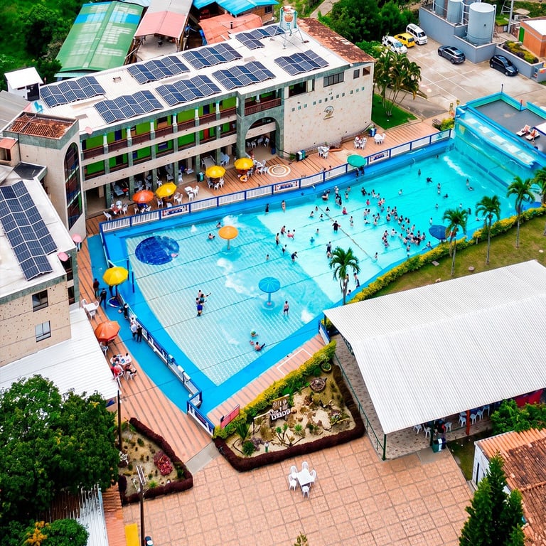Vista aérea de la piscina al aire libre con agua azul, edificación con paneles solares, zonas de descanso y visitantes disfrutando