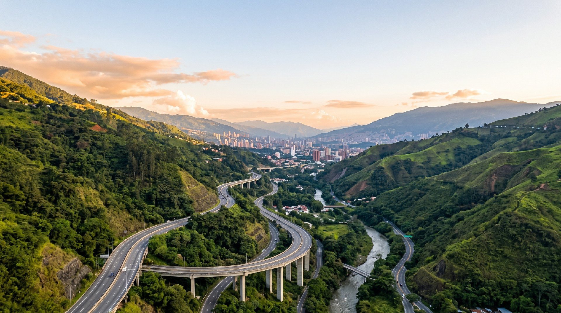 Vista aérea de un puente curvo de autopista entre montañas verdes con un río serpenteando el valle y el skyline de la ciudad al atardecer