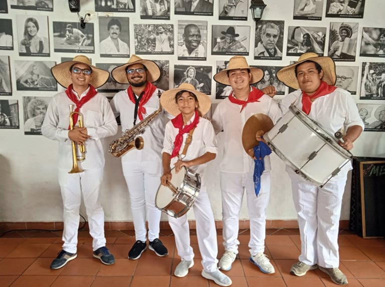 Five musicians in white outfits and straw hats with red scarves hold brass and percussion instruments against a wall of vintage photographs.