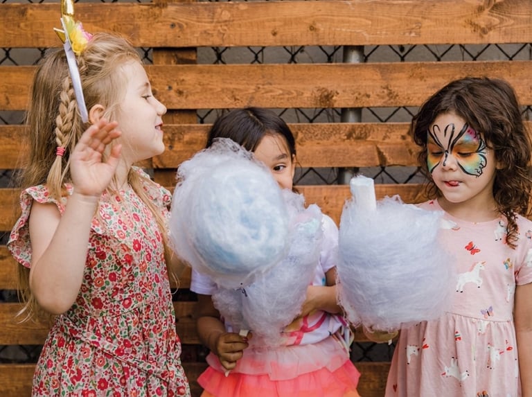 Three children enjoying cotton candy at an event, with one girl having face paint and wooden wall backdrop