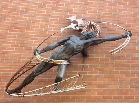 Bronze sculpture of a dynamic male figure with outstretched arms holding a whip against a red brick wall