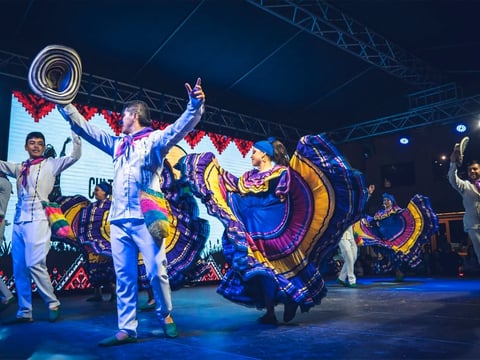 Performers in colorful traditional Mexican dance costumes on stage with vibrant blue and gold clothing during a cultural performance.