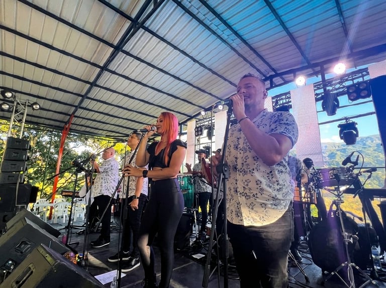 Live band performance on outdoor stage with vocalist singing into microphone, guitarist, and drummer under metal roof structure