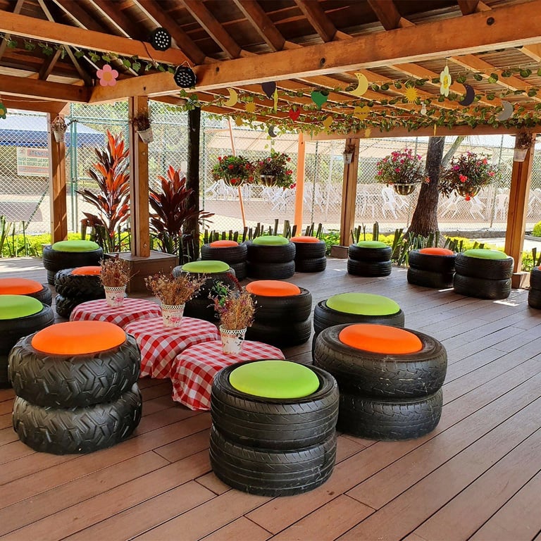 Zona de descanso cubierta con puff es de llanta y cojines naranja y verde lima bajo una pérgola de madera con flores colgantes